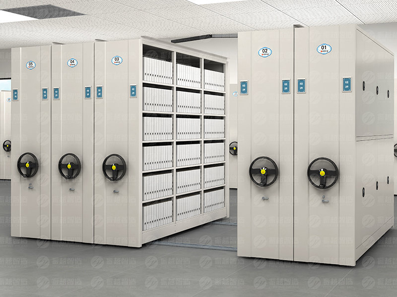 Light grey manual mobile shelving system with large black circular hand wheels featuring yellow centers, blue oval unit identification labels, and standard single-section side panel design for high-density archive storage with manual operation.
